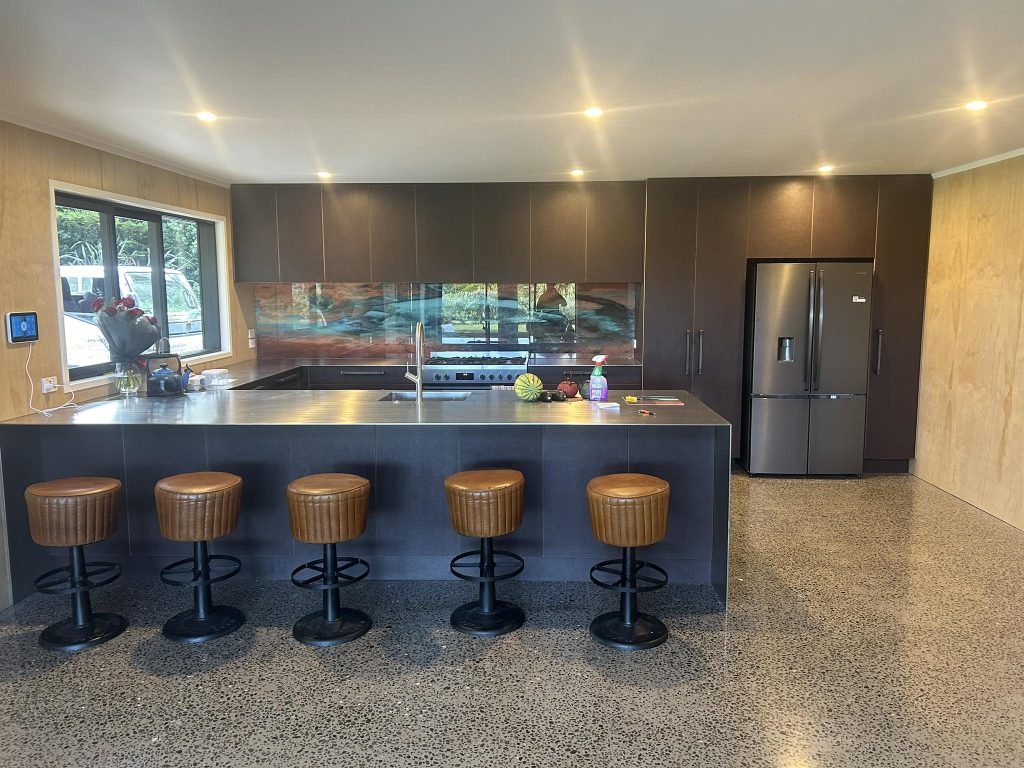 Dark modern kitchen with stainless-steel benchtop, feature glass splashback, matte cabinetry, and polished concrete flooring crafted by Newton Gordge Joinery.